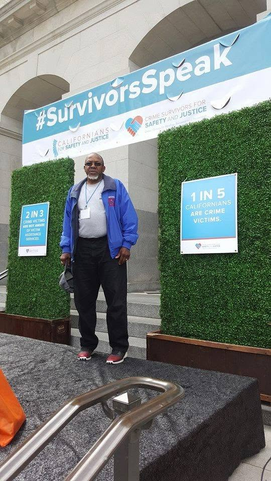 Lawrence C. Gray Jr. at the #SurvivorsSpeak rally in San Francisco, standing between signs reading '1 IN 5' about crime victims